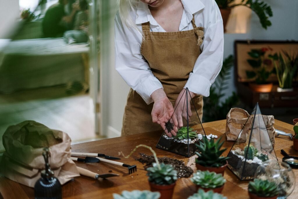 A woman in an apron assembling a beautiful indoor succulent terrarium with gardening tools.